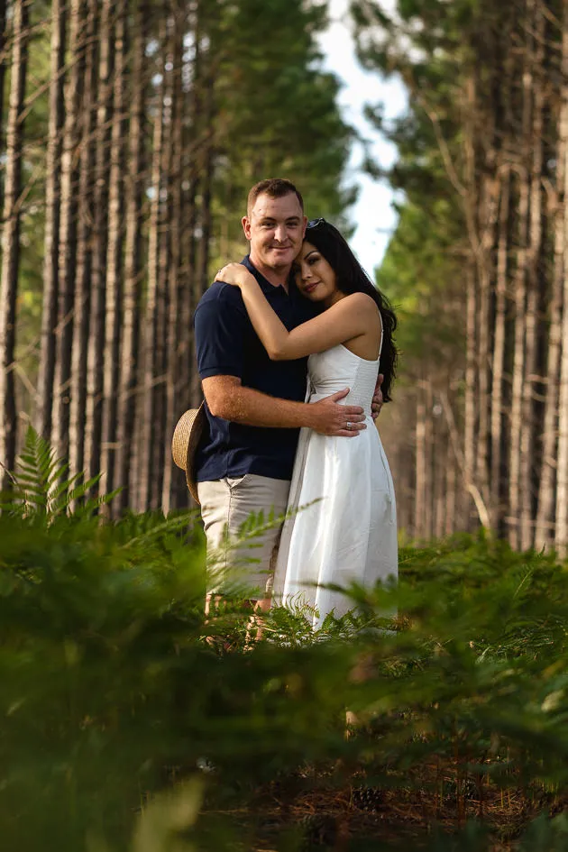 Romarie and Jake embracing among the ferns between tall rows of pines