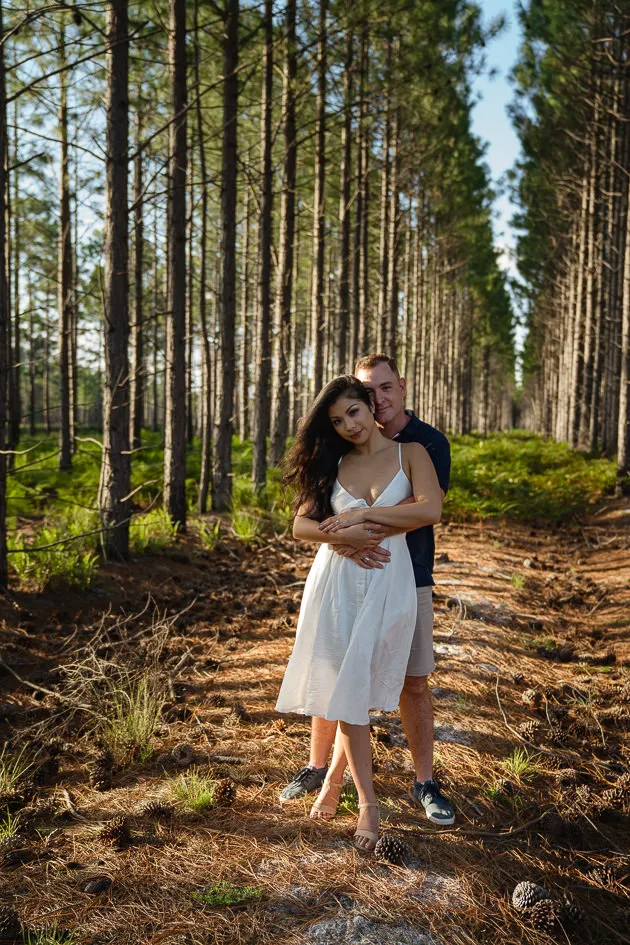 Romarie and Jake standing together on a needle-strewn path between the pines