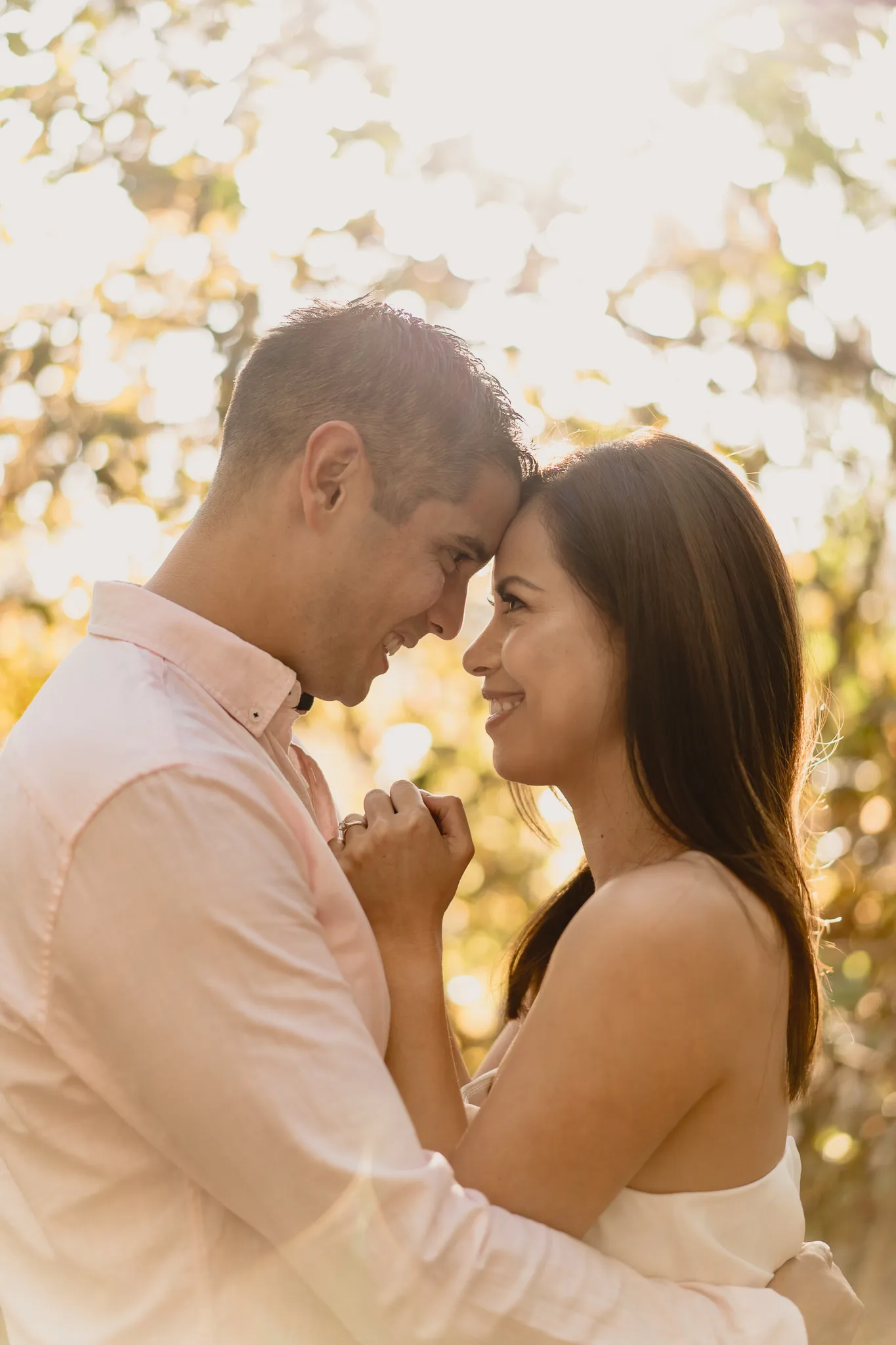 Rafaela and Mark forehead to forehead, the late sun glowing through the leaves behind them