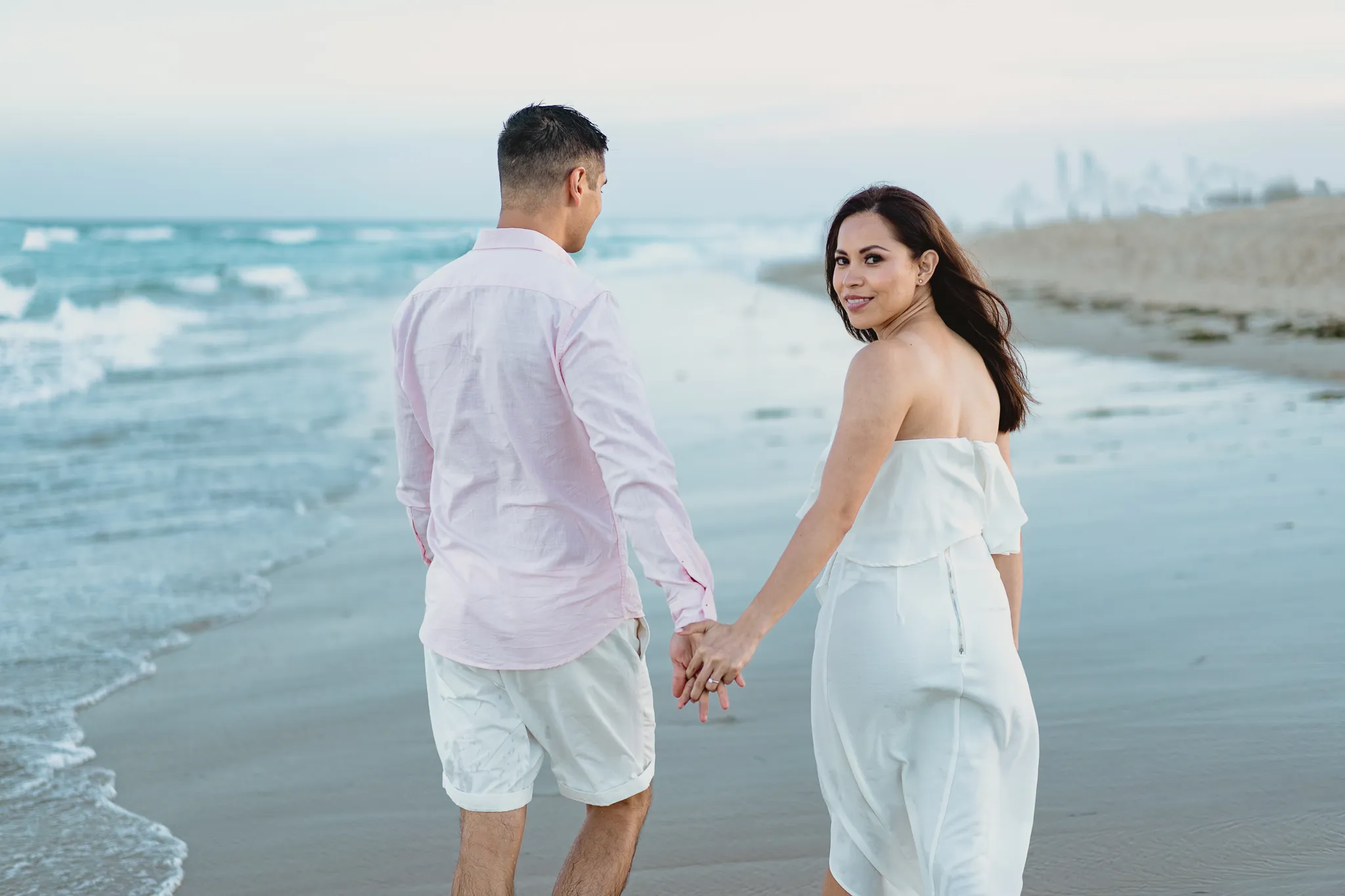 Rafaela and Mark walking along the shore hand in hand, Rafaela looking back over a shoulder