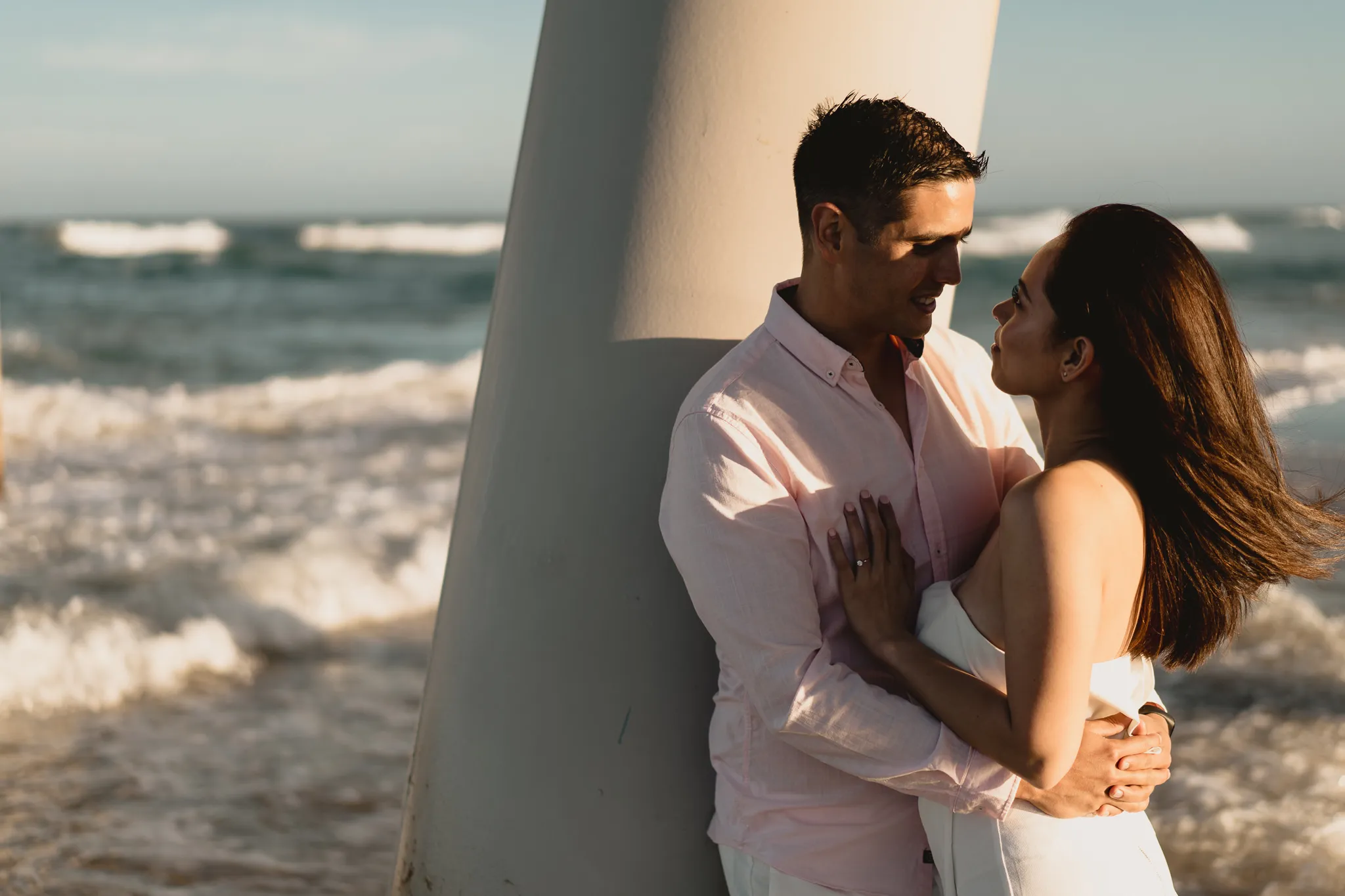 Rafaela and Mark close against a pier pylon, the surf breaking behind them