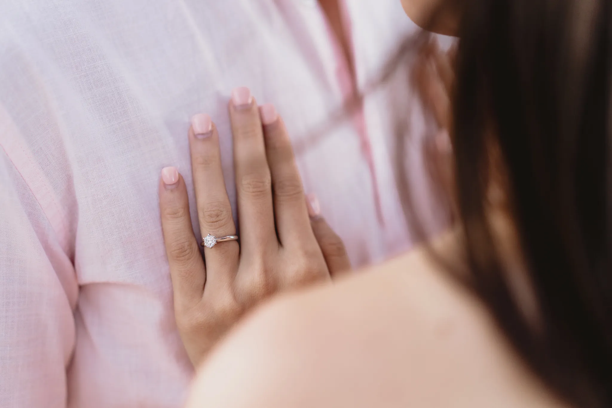 An engagement ring on a hand resting against a pale pink linen shirt