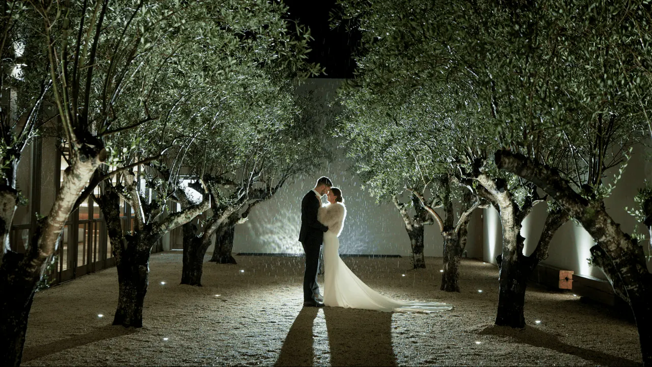 Jessica and Lachlan embracing at night at the end of a path of lit olive trees
