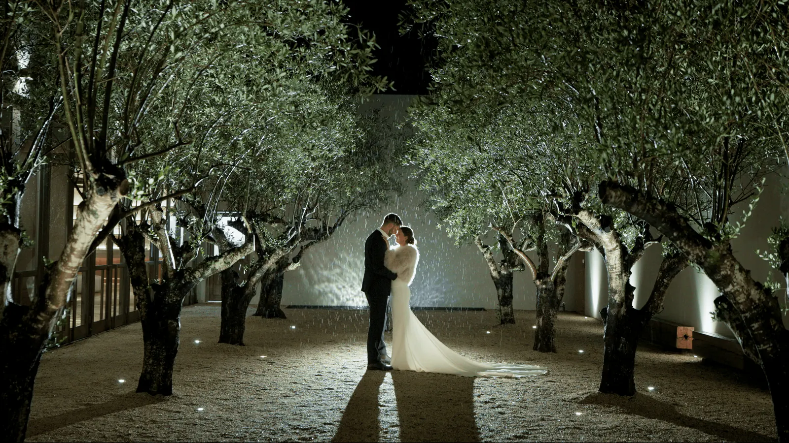 Jessica and Lachlan embracing at night at the end of a path of lit olive trees